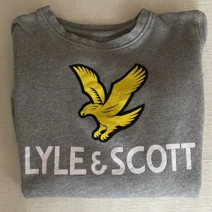 Grå sweatshirt från Lyle & Scott - Säljer en snygg grå sweatshirt från Lyle & Scott med deras ikoniska gula örnlogga på bröstet. Tröjan är i mycket bra skick och har långa ärmar. Perfekt för en avslappnad stil eller för att hålla sig varm under kyligare dagar.