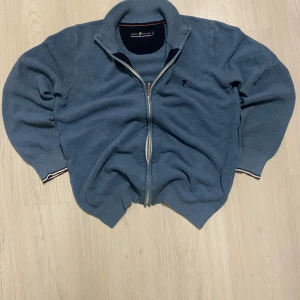 Blå stickad tröja från Denim Culture - Säljer en snygg blå stickad tröja från Denim Culture med dragkedja och broderad detalj på bröstet. Tröjan har en hög krage och ribbade muddar vid ärmslut och nederkant. Perfekt för kyliga dagar! Priset kan diskuteras!