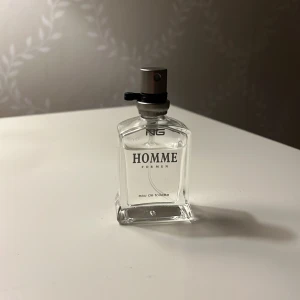 Homme Eau de Toilette för män - Elegant och stilren flaska med NG Homme Eau de Toilette. Den transparenta glasflaskan har en modern design med svart spraymunstycke och texten 'Homme' i svart. Perfekt för den som söker en klassisk och fräsch doft.