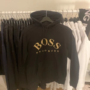 Svart hoodie från Hugo Boss - Snygg svart hoodie från Hugo Boss med stor guldfärgad logga på bröstet. Tröjan har en klassisk design med huva och dragsko samt en praktisk magficka. Perfekt för en stilren och bekväm look.