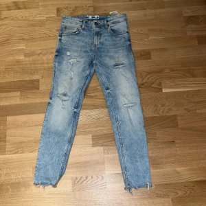 Blå slitna jeans - Säljer ett par blå slitna jeans i storlek 27/30. De har en cool distressed look med flera revor och slitningar, perfekt för en avslappnad stil. Jeansen är i bra skick och har en klassisk femficksdesign med knapp och dragkedja framtill. Passar perfekt till både vardag och fest!