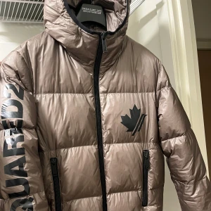 Khaki pufferjacka från Dsquared2 - Säljer en knappt använd khaki pufferjacka från Dsquared2 i storlek 48. Jackan har en logga på bröstet och ärmarna, med dragkedja och huva. Perfekt för kalla dagar med sitt äkta dunfoder. Nypris 8 995 kr!🧥