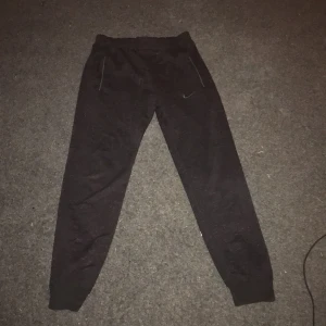 Svarta mjukisbyxor från Nike - Säljer ett par svarta mjukisbyxor från Nike med elastisk midja och dragkedjefickor. Perfekta för en avslappnad stil eller träning. Byxorna har en liten skada på tyget.