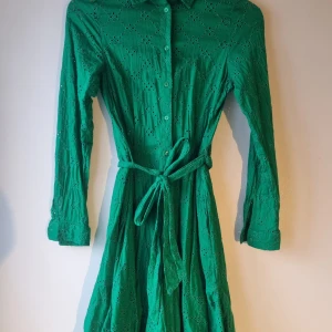 Grön klänning från Holly & Whyte - Snygg grön klänning från Holly & Whyte med broderade detaljer och knytband i midjan. Klänningen har långa ärmar och knappar framtill, perfekt för vår och sommar.