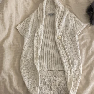 Vit stickad kofta - Säljer en supermysig vit stickad kofta med korta ärmar och en stor knapp framtill. Perfekt för lager-på-lager under hösten eller vintern. Den har ett snyggt flätmönster på sidorna och en skön, lös passform. Passar perfekt till både vardag och mysiga hemmakvällar. 🧶✨
