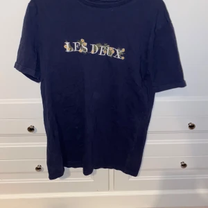 Mörkblå t-shirt från Les Deux - Säljer en mörkblå t-shirt från Les Deux med ett färgglatt tryck på bröstet. T-shirten har en rund hals och korta ärmar. Perfekt för en avslappnad stil.