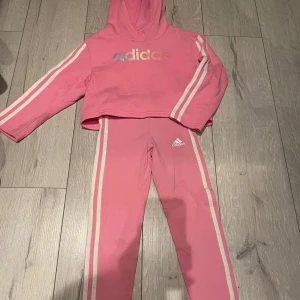 Rosa hoodie och byxor från Adidas - Säljer ett set med rosa hoodie och matchande byxor från Adidas. Hoodien har en croppad design med långa ärmar och Adidas-loggan i regnbågsfärger på bröstet. Byxorna har vita ränder längs sidorna och en liten Adidas-logga på benet. Perfekt för en sportig look! Storlek 6-7 år
