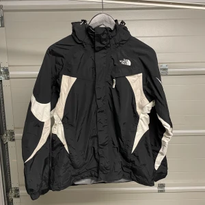 Svart och vit jacka från The North Face - Säljer en svart och vit jacka från The North Face i bra skick. Jackan har en dragkedja framtill och en justerbar huva. Perfekt för kyliga dagar och utomhusaktiviteter. Den har en sportig look med vita detaljer som ger en snygg kontrast. Säljes i befintligt skick. Storlek: M