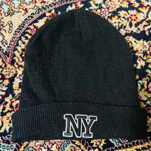Svart ribbad mössa från H&M - Snygg svart ribbad mössa från H&M med broderad 'NY' text framtill. Perfekt för att hålla värmen under kalla dagar. Mössan är i ett mjukt och stretchigt material som passar de flesta huvudstorlekar. En stilren accessoar för både vardag och fritid.