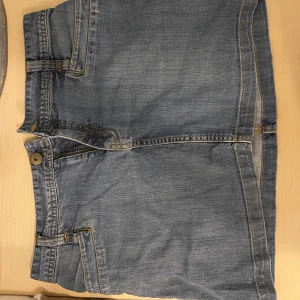 Blå jeanskjol - Snygg blå jeanskjol med klassisk design och fickor både fram och bak. Kjolen har en knapp och dragkedja framtill samt bälteshällor för extra stil. Perfekt för en avslappnad look. Gammal kollektion ifrån H&M