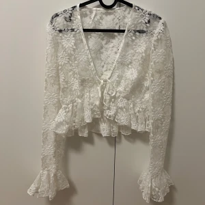 Vit spetsblus med volangdetaljer - Jättefin vit spetsblus med v-ringning och volangdetaljer. Blusen har långa ärmar och knytning framtill, står ingen storlek men passar mig som har S och kan även passar till XS