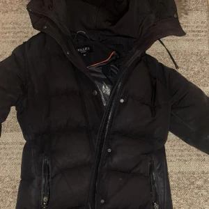 Jacka från Hollies - Säljer en svart dunjacka från Hollies med avtagbar äkta päls på huvan. Jackan har dragkedja och knappar framtill samt praktiska fickor med dragkedja. Perfekt för kalla vinterdagar.