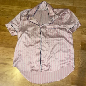 Rosa randig skjorta från Victoria's Secret - Säljer en superfin rosa randig skjorta från Victoria's Secret. Den är kortärmad och har en klassisk krage med knappar framtill. Materialet känns lyxigt och mjukt, perfekt för en avslappnad men stilren look. Pyjamas tröja