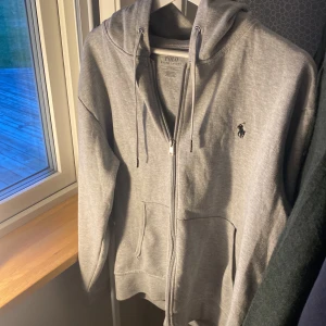 Grå hoodie från Polo Ralph Lauren - Säljer en snygg och bekväm grå hoodie från Polo Ralph Lauren. Den har en dragkedja framtill och en klassisk logga på bröstet. Perfekt för både vardag och chill. Passar bra till jeans eller joggers. 🐴