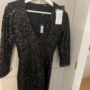 Svart paljett klänning från NLY One - Säljer en superfin svart glittrig klänning från NLY One i storlek 36. Den har en v-ringad design och trekvartsärmar, perfekt för festliga tillfällen. Klänningen är helt ny med taggar kvar. Perfekt för nyår eller en glittrig utekväll! ✨