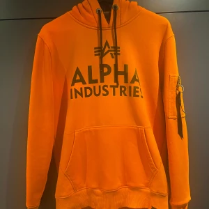 Orange hoodie från Alpha Industries - Säljer en orange hoodie från märket Alpha industries, perfekt för skidåkning bland annat då det finns en ficka vid ärmen för skipass.