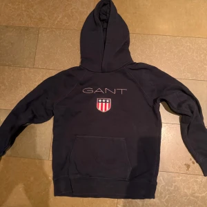 Mörkblå hoodie från GANT - Säljer en snygg mörkblå hoodie från GANT med ett emblem på bröstet. Fint skick. 