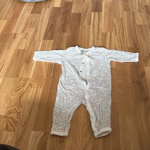 Blommig babyoverall från H&M - Söt babyoverall från H&M med blommigt mönster i rosa och grått på vit botten. Overallen har långa ärmar och knappar framtill för enkel påklädning. Perfekt för de minsta under vår och sommar.