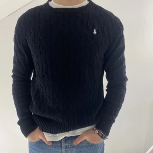 Svart kabelstickad tröja från Ralph Lauren - Säljer en stilren svart kabelstickad tröja från Polo Ralph Lauren. Tröjan har en klassisk rund halsringning och det ikoniska logotypbroderiet på bröstet. Perfekt för en elegant och avslappnad look.