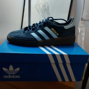 Adidas Handball Spezial  - Säljer ett par klassiska Adidas Handball Spezial sneakers i blå mocka med ljusblå ränder och texten 'Spezial' i guld. Skorna har en brun sula och snörning framtill. Perfekta för en sportig och stilren look.