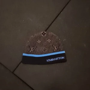  mössa från Louis Vuitton - Snygg mössa från Louis Vuitton med deras ikoniska monogrammönster. Mössan har en blå rand och märkesnamnet broderat i samma färg. Perfekt för att hålla stilen i kallare väder.