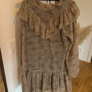 Leopardmönstrad klänning med volang - Trendig leopardmönstrad klänning med volangdetaljer. Klänningen har långa ärmar och en lös passform, perfekt för en avslappnad stil. Den är tillverkad i ett lätt och bekvämt material.