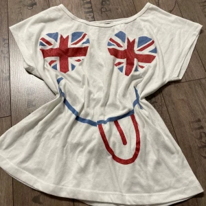 Vit t-shirt med Union Jack-motiv - Säljer en vit t-shirt med korta ärmar och ett stort smiley-tryck med Union Jack-motiv på framsidan. Baksidan har dekorativa utskärningar upptill. Perfekt för en avslappnad stil.