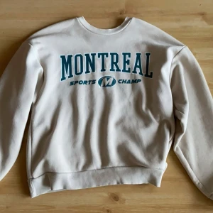 Beige sweatshirt från Gina Tricot - Säljer en beige sweatshirt från Gina Tricot med texten 'Montreal Sports Champ' i grönt på framsidan. Tröjan har en rund halsringning och ribbade muddar vid ärmslut och nederkant. Perfekt för en avslappnad stil.