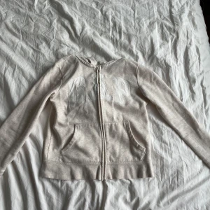 Beige hoodie från GAP Kids - Säljer en beige hoodie från GAP Kids med dragkedja och fickor framtill. Tröjan har en stor GAP-logga på bröstet och en bekväm huva. Perfekt för en avslappnad stil.