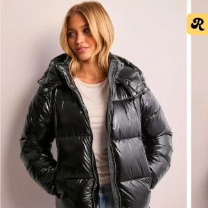 Svart glansig pufferjacka - Snygg svart glansig pufferjacka med dragkedja och huva. Perfekt för kyliga dagar med sin vadderade design som håller dig varm. Jackan har en modern look och passar bra till jeans eller leggings.