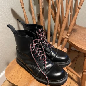 Doc Martens Svarta boots med snörning - Säljer ett par klassiska svarta Dr. Martens boots med röd- och vitmönstrade snören. Skorna är tillverkade i skinn och har en robust design med dragflik baktill för enkel påtagning. Storlek 47. Knappt använda 