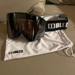 Skidglasögon Bliz - Snygga svarta skidglasögon från Bliz med bred elastisk rem och spegelglas. Perfekta för vintersport och skyddar mot starkt solljus. Kommer med en praktisk vit förvaringspåse med Bliz-logga. Nypris=999kr 