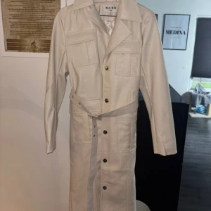 Vit trenchcoat från NA-KD - Snygg vit trenchcoat från NA-KD med knappar och bälte i midjan. Jackan har långa ärmar och flera fickor framtill. Perfekt för en stilren look.