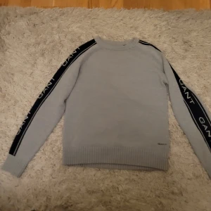 Grå tröja från GANT - Säljer en stilren grå tröja från GANT med svarta detaljer längs ärmarna där märkesnamnet är broderat. Tröjan har ribbade muddar vid ärmslut och nederkant, vilket ger en snygg passform. Perfekt för en avslappnad stil. Använd en eller två gånger.