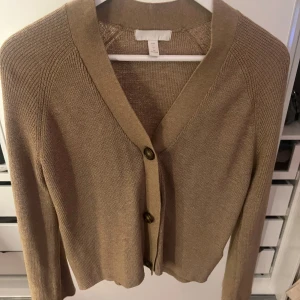 Beige kofta från H&M - Säljer en stilren beige kofta från H&M. Den har en klassisk design med knappar framtill och långa ärmar. 
