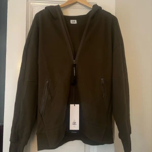 Oliv grön  hoodie från C.P. Company - Snygg olivgrön hoodie från C.P. Company , hoddie kom som en present men säljs nu för att den inte kommer till användning, tröjan är oanvänd och direkt från C.P Companys butik som köptes för 3299kr