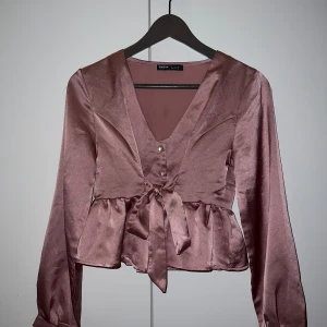 Rosa satinblus  - Elegant rosa satinblus med v-ringning och knytband i midjan. Blusen har långa ärmar och en fin volangdetalj nedtill. Perfekt för en stilfull look.