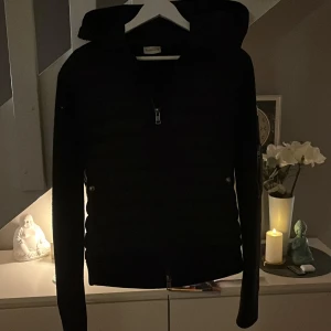 Svart dunjacka från Moncler - Säljer en stilren svart dunjacka från Moncler med huva och dragkedja. Perfekt för kyliga dagar med sin quiltade design som ger extra värme. Jackan har en klassisk passform och är ett måste för garderoben.