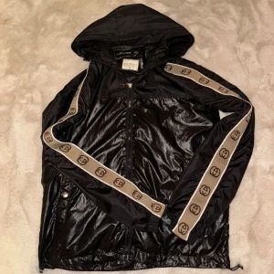 Svart vindjacka från Gucci - Snygg svart vindjacka från Gucci med huva och dragkedja. Jackan har dekorativa band med logotyp längs ärmarna och tryckknappar på fickorna. Perfekt för en stilren look.