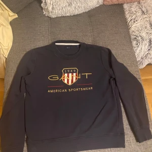 Svart sweatshirt från GANT - Säljer en stilren svart sweatshirt från GANT med tryck i gult och rött. Tröjan har långa ärmar, perfekt för en avslappnad look. GANT-loggan och texten 'American Sportswear' pryder framsidan.