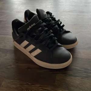 Svarta sneakers från Adidas - Snygga svarta Adidas sneakers med vita detaljer och klassiska tre ränder på sidan. Skorna har snörning och en vit sula som ger en stilren look. Perfekta för en avslappnad stil.