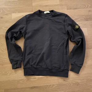 Mörkblå sweatshirt från Stone Island - Säljer en stilren mörkblå sweatshirt från Stone Island med deras ikoniska märke på ärmen. Tröjan har en klassisk rund hals och långa ärmar, perfekt för en avslappnad look. 