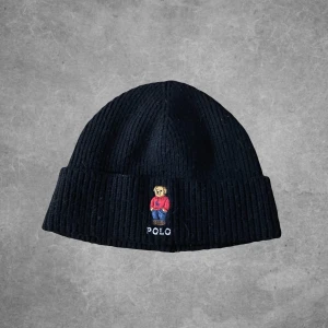 Svart ribbad mössa från Polo Ralph Lauren - Snygg svart ribbad mössa från Polo Ralph Lauren med ett broderat motiv av en björn i färgglada kläder. Perfekt för att hålla värmen med stil.