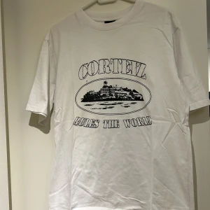 Vit t-shirt med tryck från Corteiz - Säljer en vit t-shirt från Corteiz med ett stort svart tryck på framsidan som föreställer en byggnad och texten 'Corteiz Rules The World'. T-shirten har en klassisk passform och är kortärmad. Perfekt för en avslappnad stil.