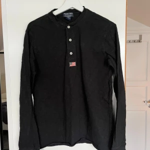 Svart långärmad tröja från Ralph Lauren - Säljer en stilren svart långärmad tröja från Ralph Lauren med knappar framtill och ett litet märke på bröstet. Perfekt för en avslappnad look. Tillverkad i 100% bomull för extra komfort.