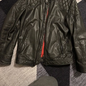 Svart skinnjacka med quiltade detaljer - Snygg svart skinnjacka från Infinity med quiltade axelpartier och röd innerfoder. Jackan har dragkedja framtill och två fickor med dragkedjor. Perfekt för en cool och stilren look.