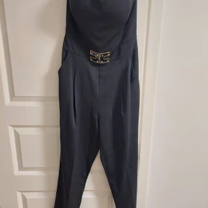 Svart jumpsuit från Aynan - Elegant svart jumpsuit från Aynan med en stilren design och ett dekorativt bälte i midjan. Perfekt för en chic look.