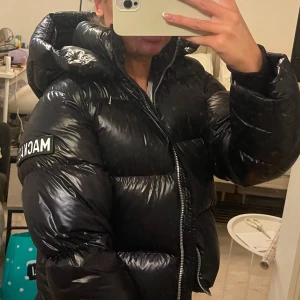 Svart pufferjacka från Mackage - Snygg svart pufferjacka från Mackage med glansigt material och dragkedja framtill. Jackan har en stor huva och långa ärmar, perfekt för kyliga dagar. Märkeslogga synlig på ärmen.