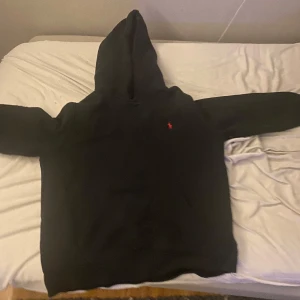 Svart hoodie från Ralph Lauren - Säljer en klassisk svart hoodie från Ralph Lauren med en liten röd logga på bröstet. Perfekt för en avslappnad stil med en bekväm passform och huva.
