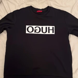 Svart tröja från Hugo Boss - Snygg svart tröja från Hugo Boss med vit logga på framsidan. Tröjan har rund hals och långa ärmar.